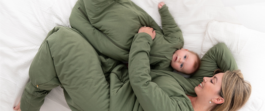 Eine Mutter und ihr Baby liegen nebeneinander auf einem Bett. Beide tragen passende grüne Schlafsäcke und lächeln. Die Mutter hat ihren Arm um das Baby gelegt, das sie liebevoll ansieht.