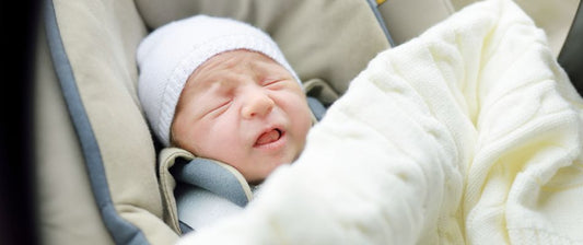 Ein neugeborenes Baby sitzt sicher in einem Autositz und ist in eine warme Decke eingewickelt. Es hat eine weiße Mütze auf.
