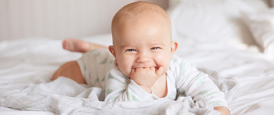 Ein lächelndes Baby liegt auf dem Bauch und steckt seine Hand in den Mund, während es auf einem Bett liegt.