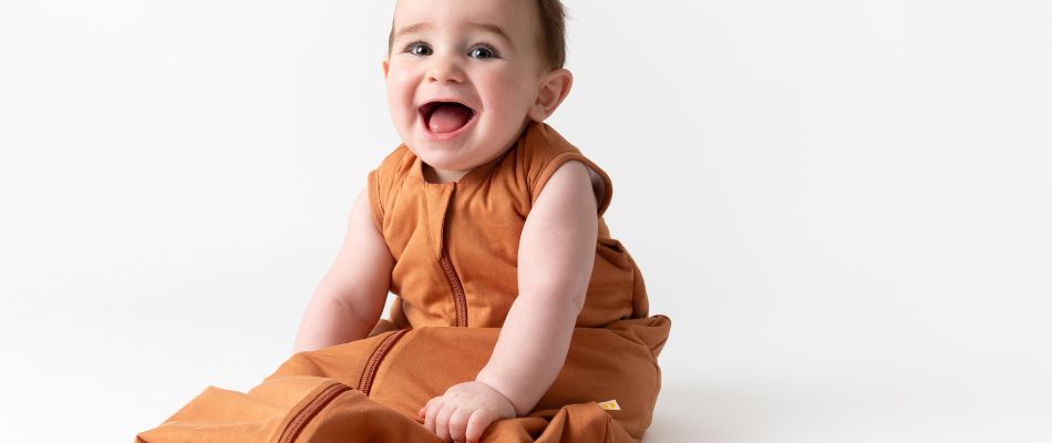 Ein glückliches Baby in einem orangefarbenen Strampler sitzt und lacht auf einem weißen Hintergrund.