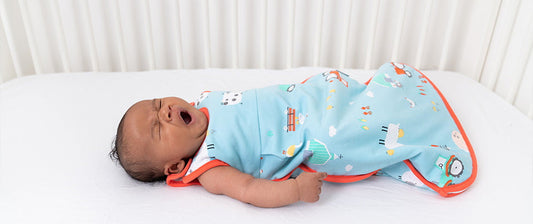 Ein Baby liegt in einem farbenfrohen Babyschlafsack auf einem weißen Bett und gähnt.