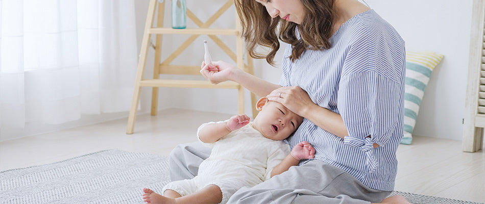 Eine Mutter sitzt auf dem Boden und hält ihr krankes Baby im Arm. Sie hat ein Thermometer in der Hand und überprüft die Temperatur des Babys, indem sie eine Hand an dessen Stirn legt.