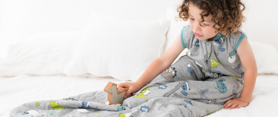 Ein Kind mit lockigem Haar sitzt auf einem Bett und spielt mit einem kleinen Holzspielzeug. Es trägt einen grauen Schlafsack mit bunten Dinosauriermotiven. Das Bett ist mit weißen Kissen und Bettwäsche ausgestattet.