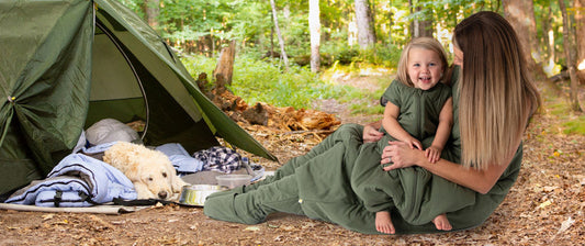 Eine Mutter und ihr Kind sitzen in grünen Schlafsäcken mit Beinen auf dem Boden eines Waldgebiets, neben ihnen steht ein grünes Zelt, in dem ein Hund auf einer Decke liegt.