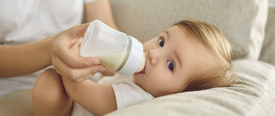 Ein Baby liegt gemütlich auf einem Sofa und trinkt aus einer Milchflasche, die von einer erwachsenen Person gehalten wird.