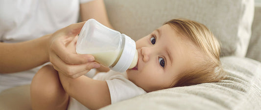 Ein Baby liegt gemütlich auf einem Sofa und trinkt aus einer Milchflasche, die von einer erwachsenen Person gehalten wird.