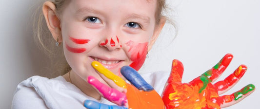 Ein lachendes Mädchen zeigt stolz ihre bemalten Hände, die in leuchtenden Farben wie Rot, Blau, Gelb und Grün gefärbt sind.