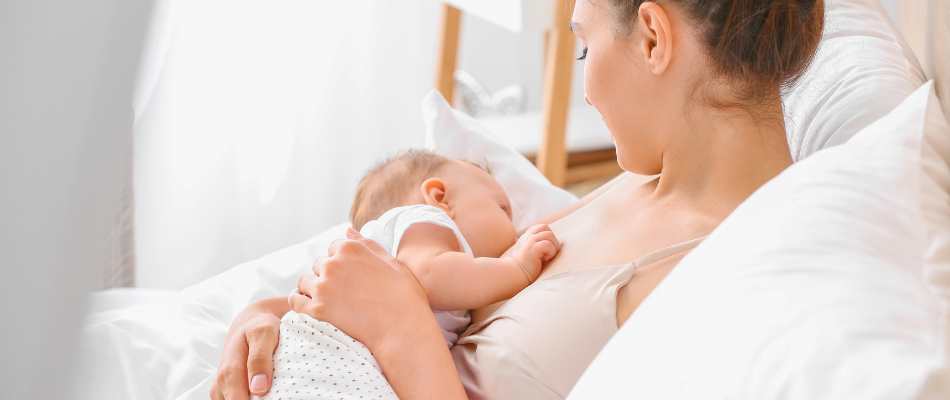 Eine Mutter sitzt in einem gemütlichen Bett und stillt ihr Baby, das in ihren Armen liegt. Sie schaut liebevoll auf ihr Kind hinunter, während sie es an ihre Brust hält.