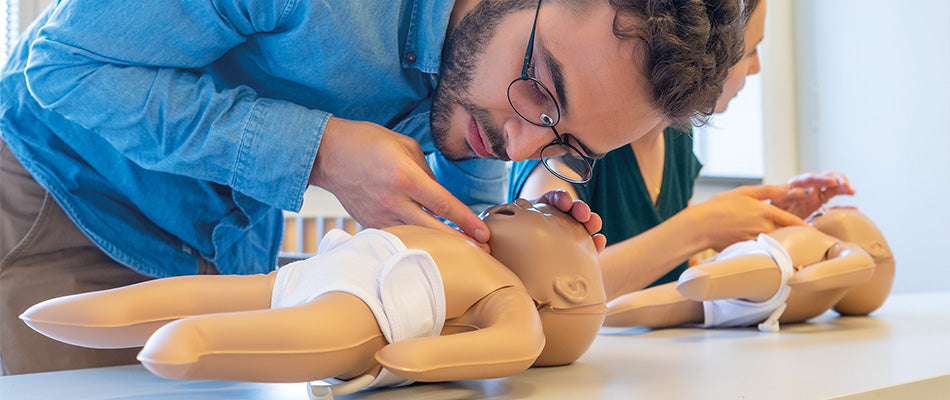 Ein Mann führt an einer Baby-Puppe eine Erste-Hilfe-Maßnahme durch, während eine Frau im Hintergrund übt.