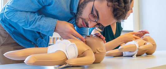 Ein Mann führt an einer Baby-Puppe eine Erste-Hilfe-Maßnahme durch, während eine Frau im Hintergrund übt.