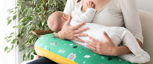 Eine Frau stillt ihr Baby, das auf einem grünen Stillkissen mit Waldmotiven liegt. Beide tragen weiße Kleidung, und im Hintergrund ist eine grüne Pflanze zu sehen