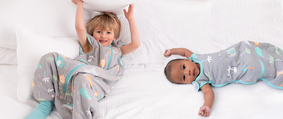 Ein Baby im Babyschlafsack und ein Junge im Schlafsack mit Füßen liegen auf dem weißen Bett