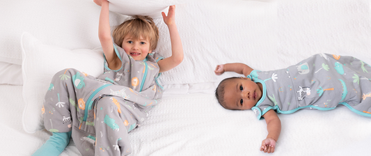 Ein Baby im Babyschlafsack und ein Junge im Schlafsack mit Füßen liegen auf dem weißen Bett