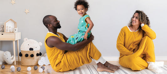 Ein Mann und eine Frau tragen passende gelbe Schlafsäcke, während das Kind in einem grünen Schlafsack mitBeinen zwischen ihnen sitzt.