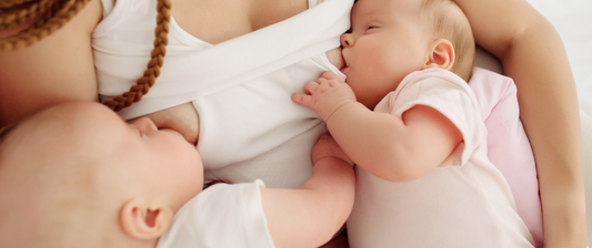 Zwei Babys werden gleichzeitig gestillt, jedes an einer Brust der Mutter. Beide Babys tragen helle Strampler und sind eng an die Mutter gekuschelt.