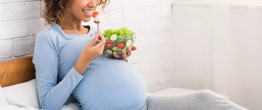 Schwangere Frau isst Salat aus einer Glasschüssel und lächelt dabei.