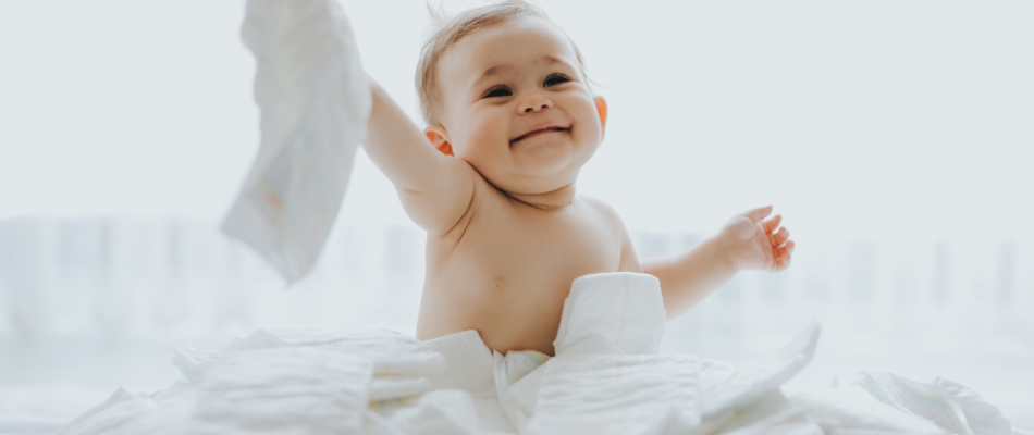 Ein fröhliches Baby sitzt umgeben von Windeln und hält eine Windel in der Hand. Es trägt keine Kleidung und lächelt.