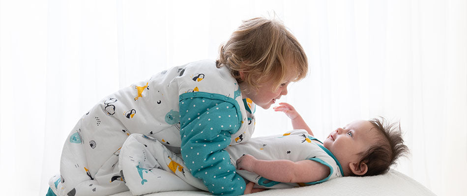 Ein Kleinkind im Schlafsack beugt sich liebevoll über ein Baby, das ebenfalls in einem Babyschlafsack liegt.